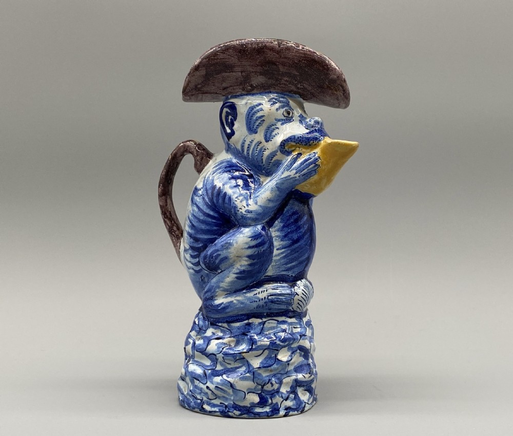 An eighteenth-century Dutch Delft tin-glazed earthenware jug shaped as a seated monkey wearing a hat and holding a vessel, decorated in blue, white, brown, and yellow glazes. View II