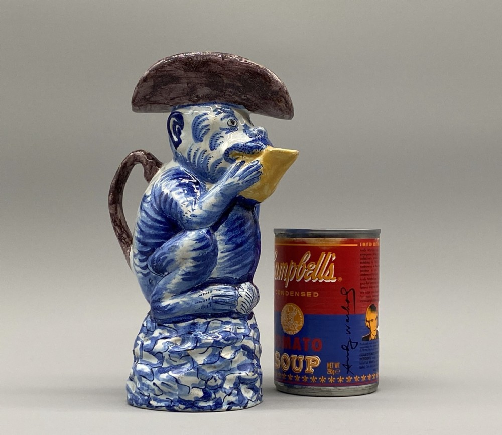 An eighteenth-century Dutch Delft tin-glazed earthenware jug shaped as a seated monkey wearing a hat and holding a vessel, decorated in blue, white, brown, and yellow glazes. View III