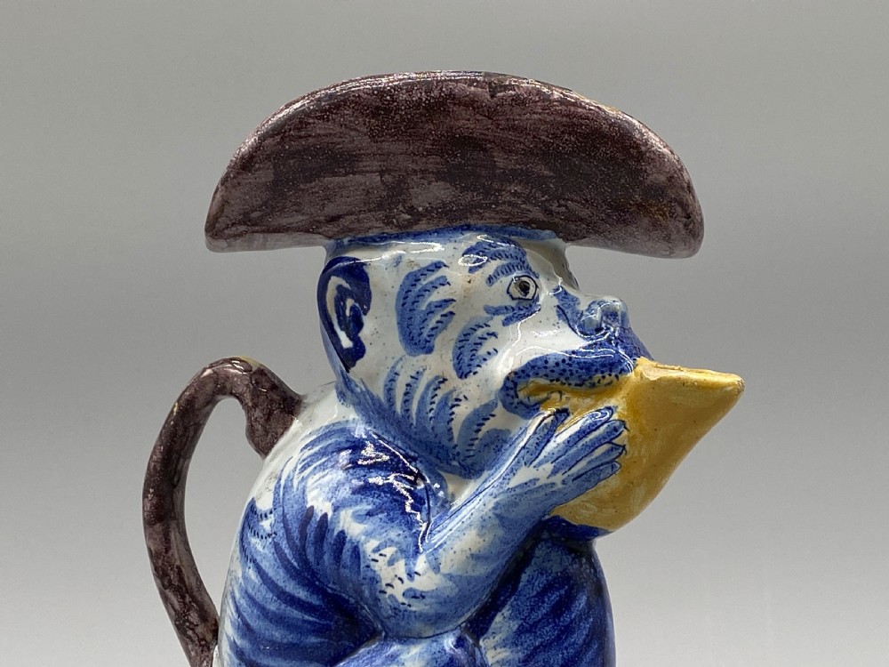 An eighteenth-century Dutch Delft tin-glazed earthenware jug shaped as a seated monkey wearing a hat and holding a vessel, decorated in blue, white, brown, and yellow glazes. View X