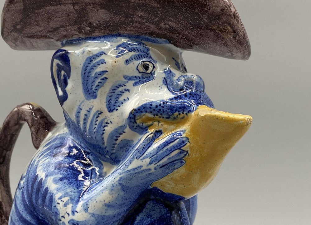 An eighteenth-century Dutch Delft tin-glazed earthenware jug shaped as a seated monkey wearing a hat and holding a vessel, decorated in blue, white, brown, and yellow glazes. View XI