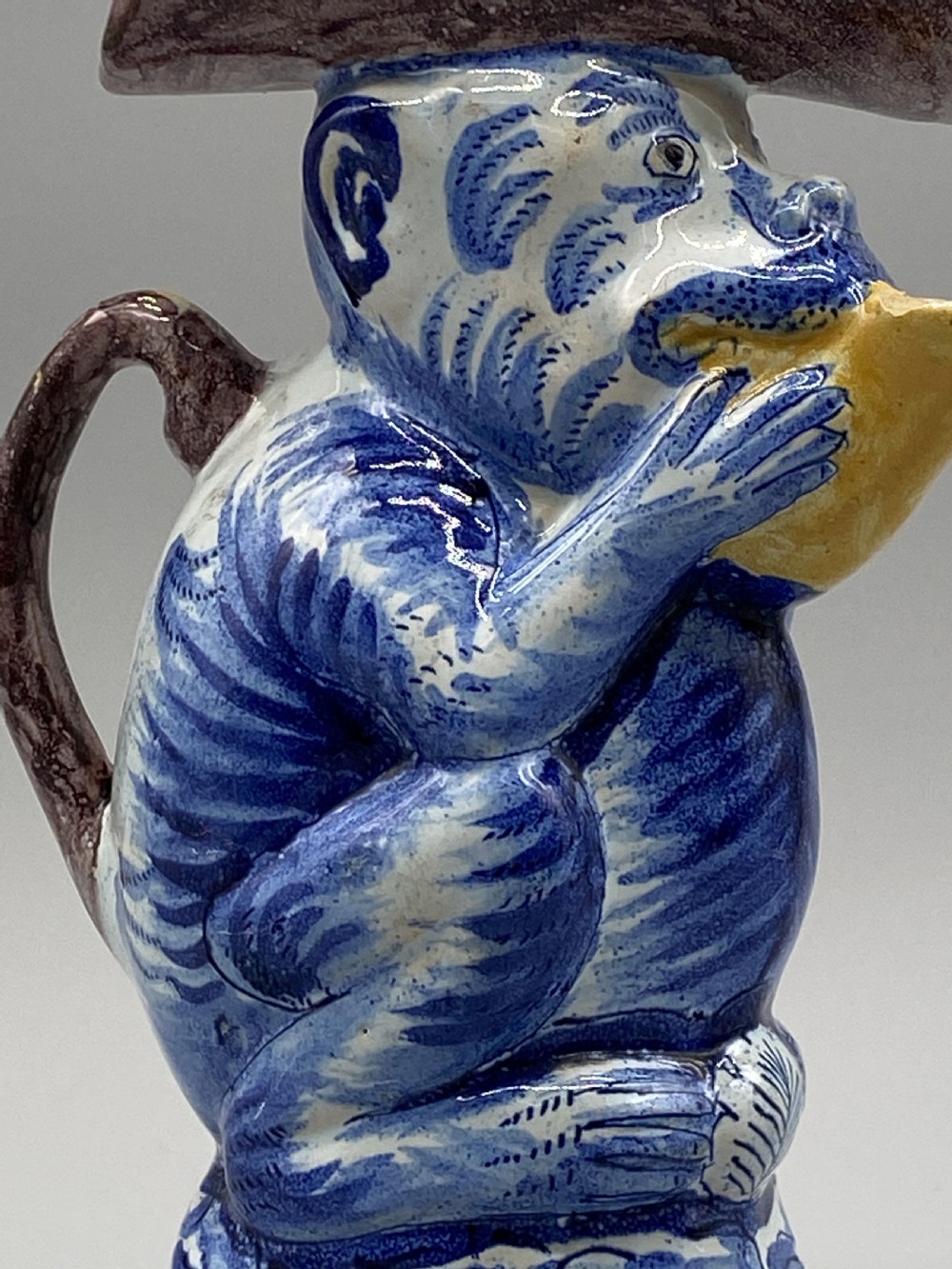 An eighteenth-century Dutch Delft tin-glazed earthenware jug shaped as a seated monkey wearing a hat and holding a vessel, decorated in blue, white, brown, and yellow glazes. View XIII