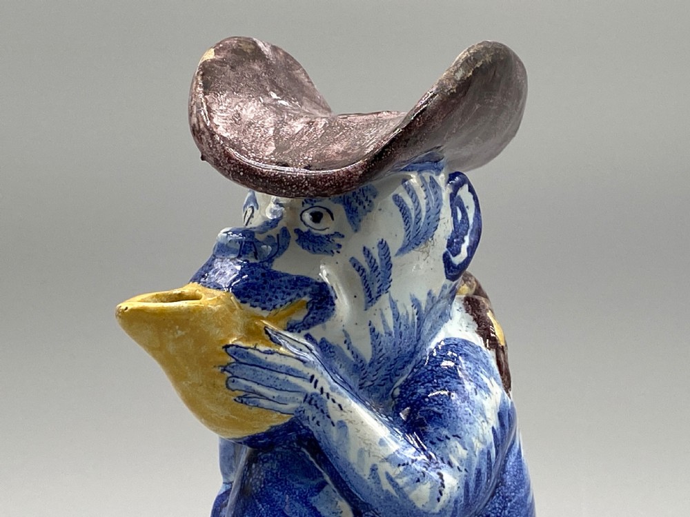 An eighteenth-century Dutch Delft tin-glazed earthenware jug shaped as a seated monkey wearing a hat and holding a vessel, decorated in blue, white, brown, and yellow glazes. View XIV