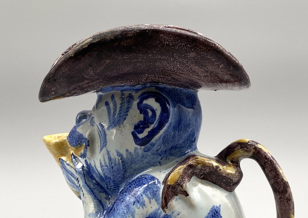 An eighteenth-century Dutch Delft tin-glazed earthenware jug shaped as a seated monkey wearing a hat and holding a vessel, decorated in blue, white, brown, and yellow glazes. View XVII