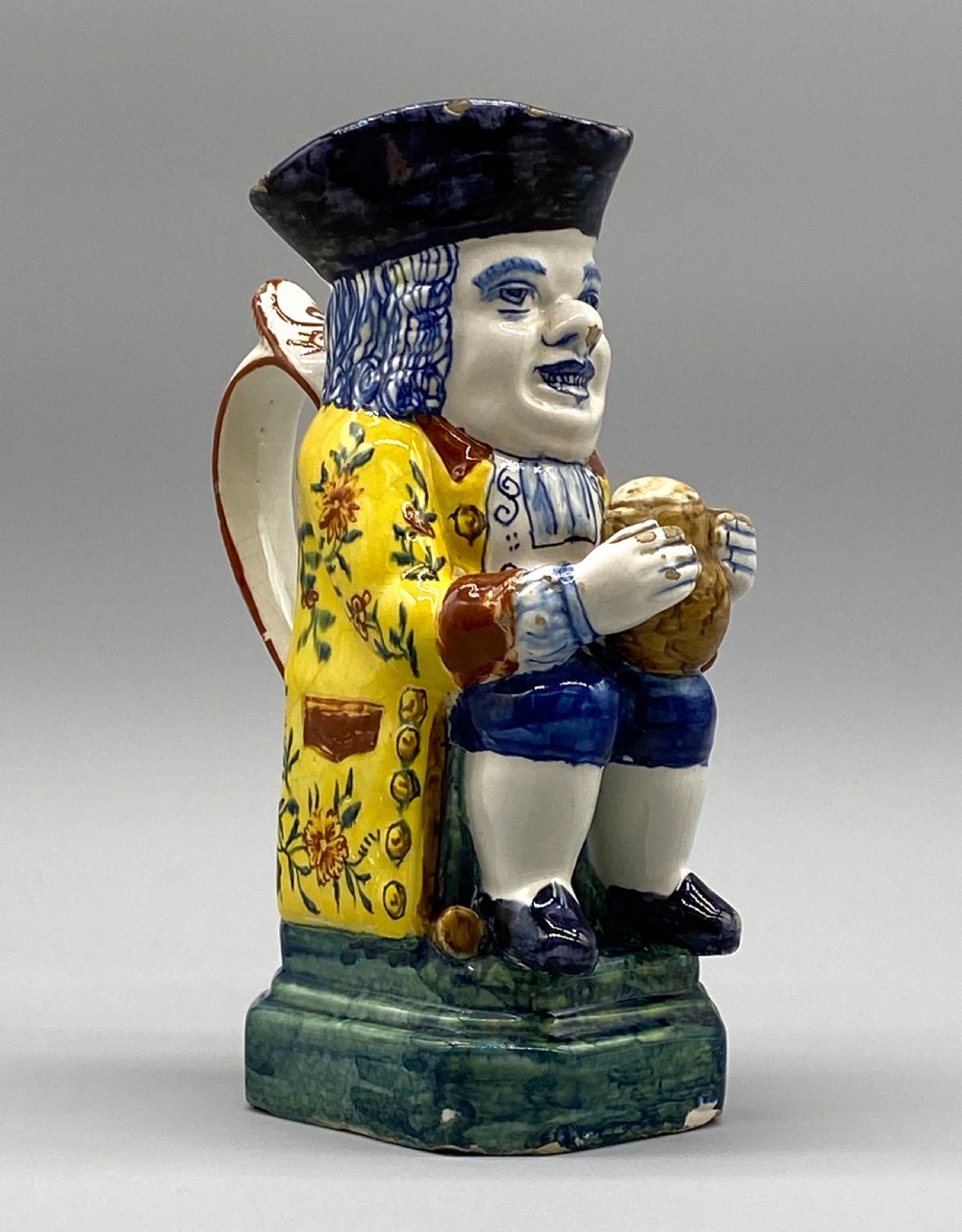 A polychrome tin-glazed earthenware Toby jug depicting a seated figure in a tricorn hat and yellow floral coat holding a small pitcher.