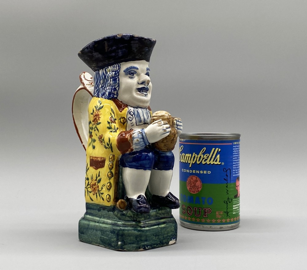 A polychrome tin-glazed earthenware Toby jug depicting a seated figure in a tricorn hat and yellow floral coat holding a small pitcher. View II