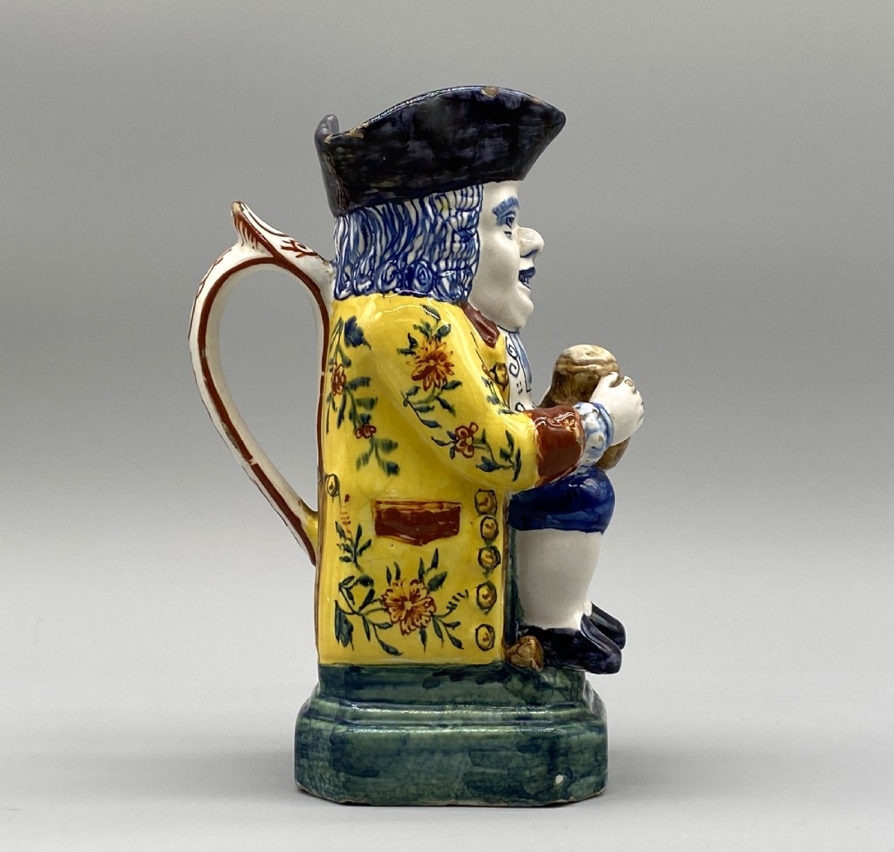 A polychrome tin-glazed earthenware Toby jug depicting a seated figure in a tricorn hat and yellow floral coat holding a small pitcher. View III