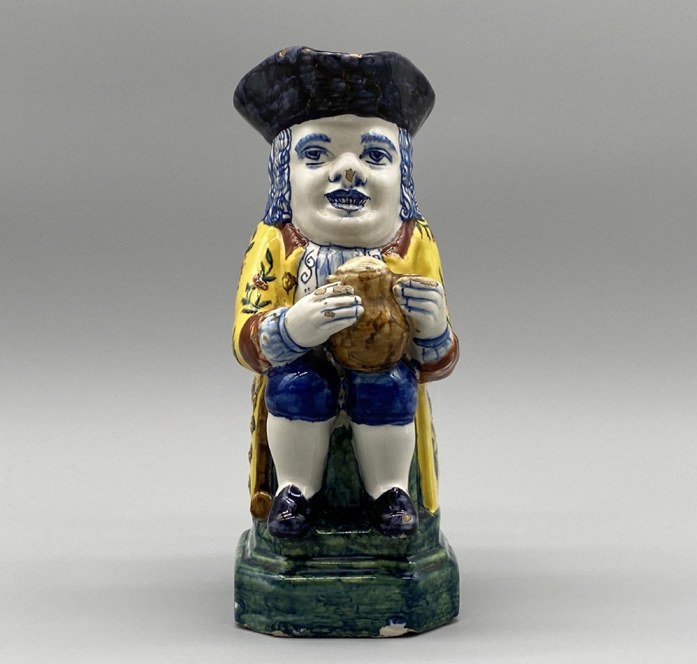 A polychrome tin-glazed earthenware Toby jug depicting a seated figure in a tricorn hat and yellow floral coat holding a small pitcher. View V