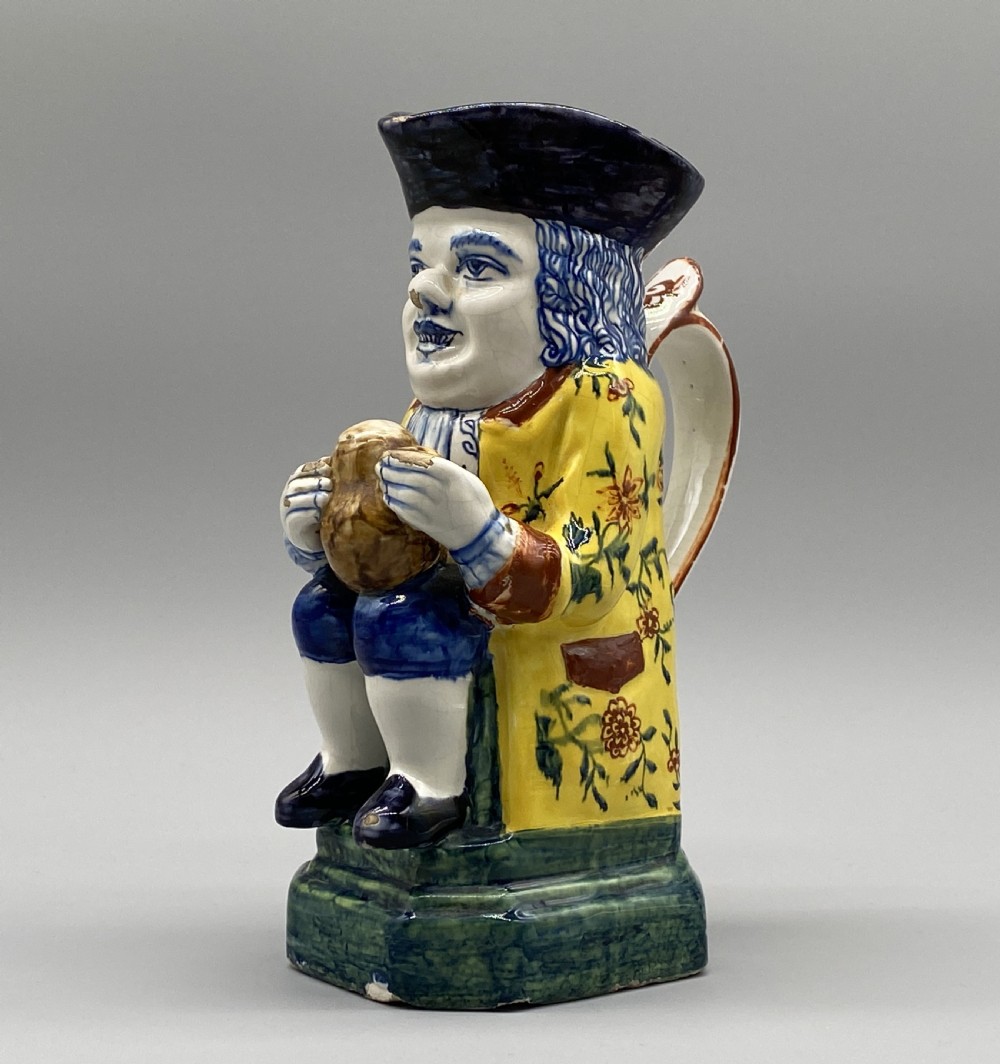 A polychrome tin-glazed earthenware Toby jug depicting a seated figure in a tricorn hat and yellow floral coat holding a small pitcher. View VI