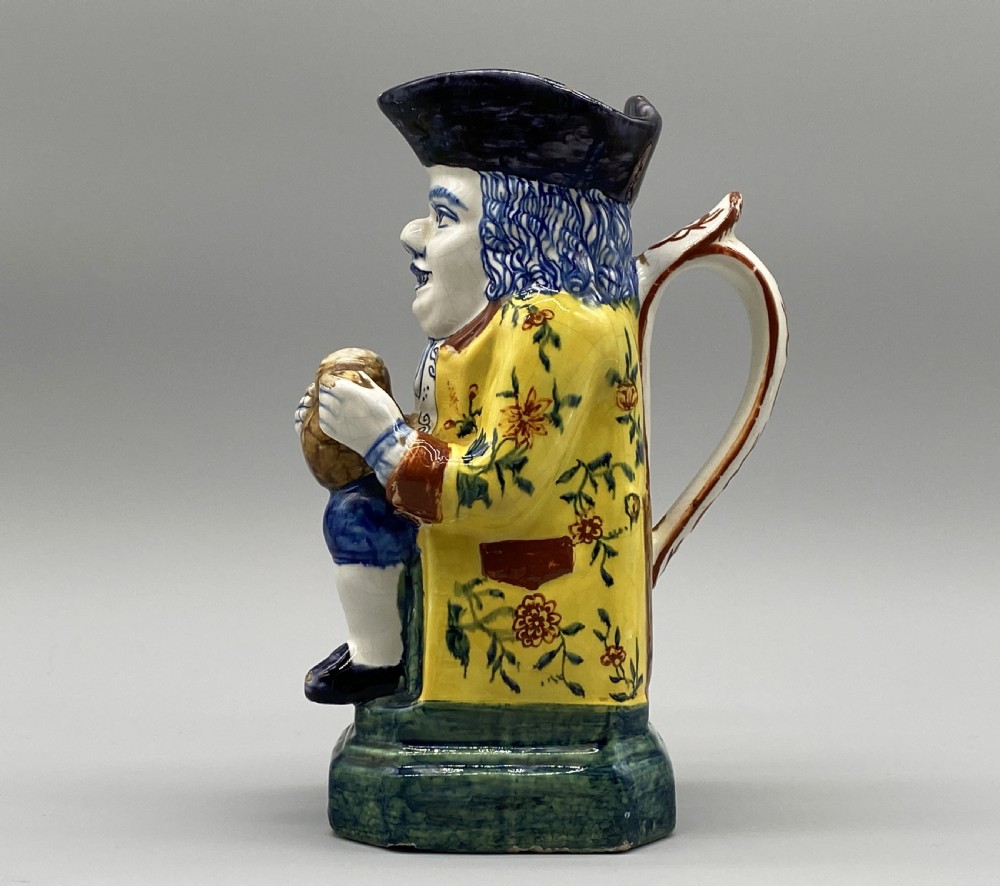 A polychrome tin-glazed earthenware Toby jug depicting a seated figure in a tricorn hat and yellow floral coat holding a small pitcher. View VII