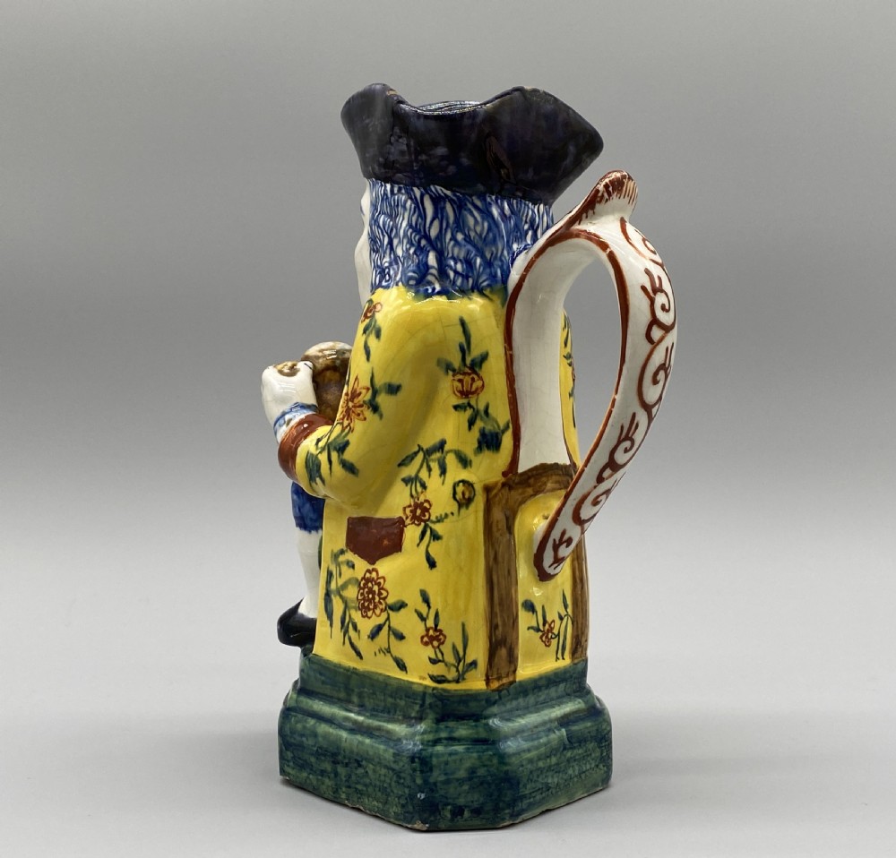 A polychrome tin-glazed earthenware Toby jug depicting a seated figure in a tricorn hat and yellow floral coat holding a small pitcher. View VIII