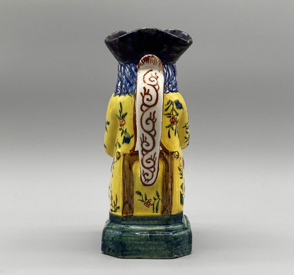 A polychrome tin-glazed earthenware Toby jug depicting a seated figure in a tricorn hat and yellow floral coat holding a small pitcher. View IX