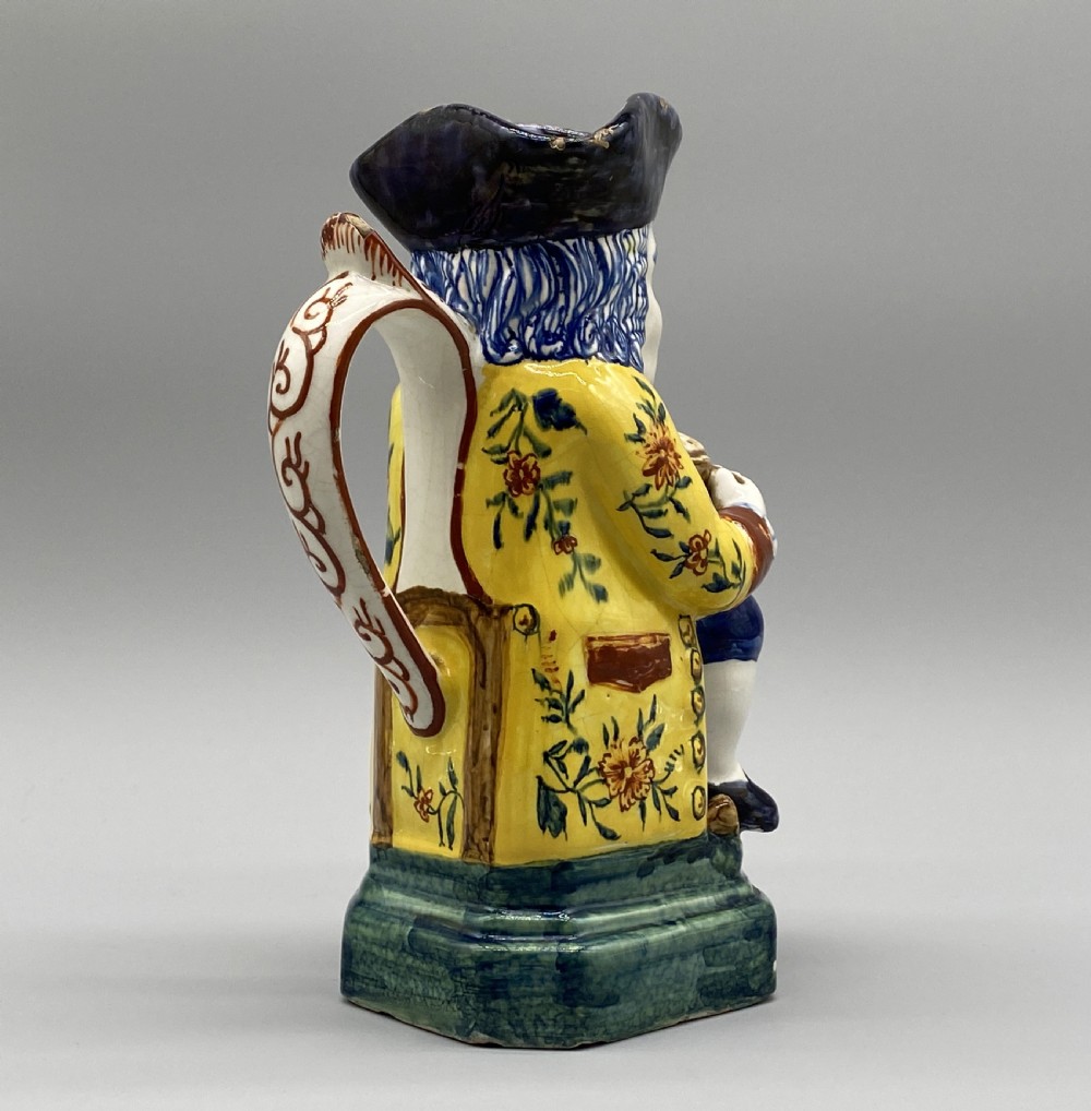 A polychrome tin-glazed earthenware Toby jug depicting a seated figure in a tricorn hat and yellow floral coat holding a small pitcher. View X