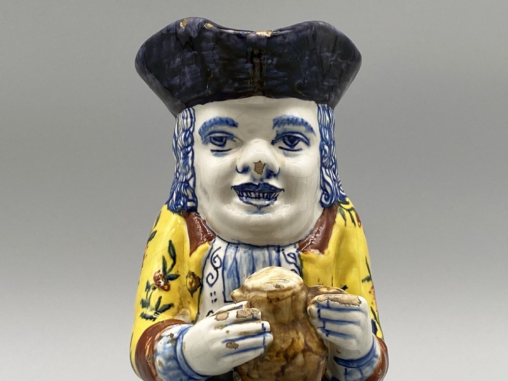 A polychrome tin-glazed earthenware Toby jug depicting a seated figure in a tricorn hat and yellow floral coat holding a small pitcher. View XI