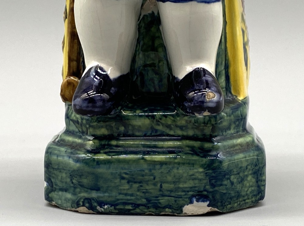 A polychrome tin-glazed earthenware Toby jug depicting a seated figure in a tricorn hat and yellow floral coat holding a small pitcher. View XIII