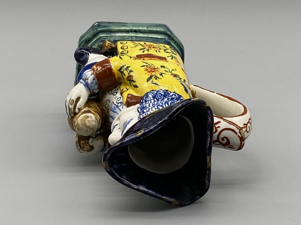 A polychrome tin-glazed earthenware Toby jug depicting a seated figure in a tricorn hat and yellow floral coat holding a small pitcher. View XIV