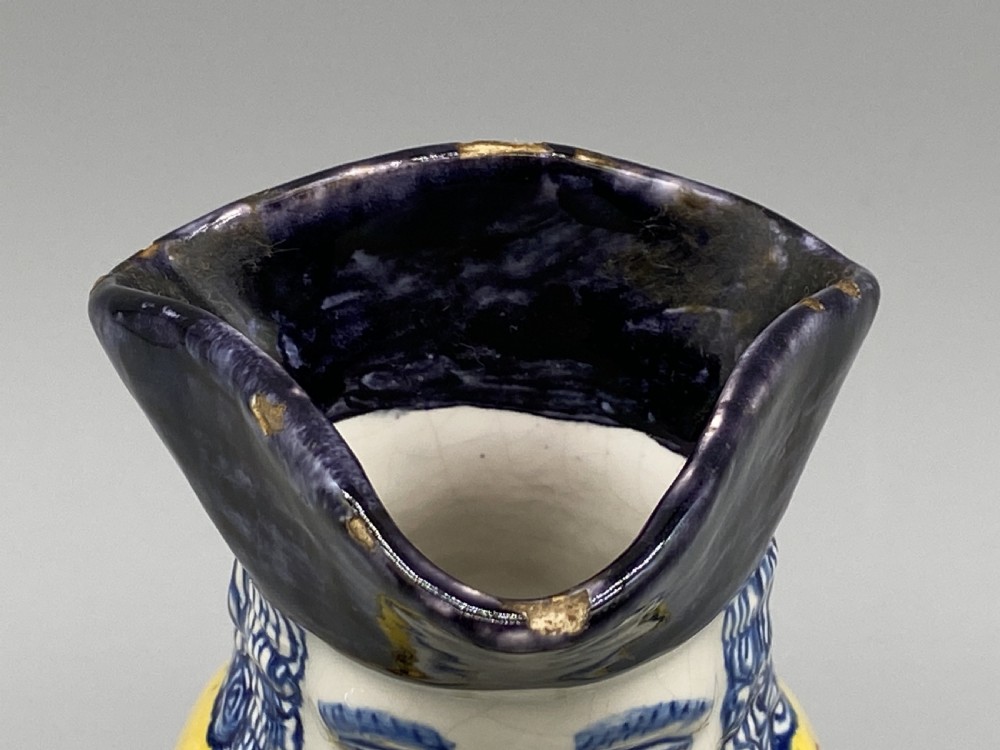 A polychrome tin-glazed earthenware Toby jug depicting a seated figure in a tricorn hat and yellow floral coat holding a small pitcher. View XV