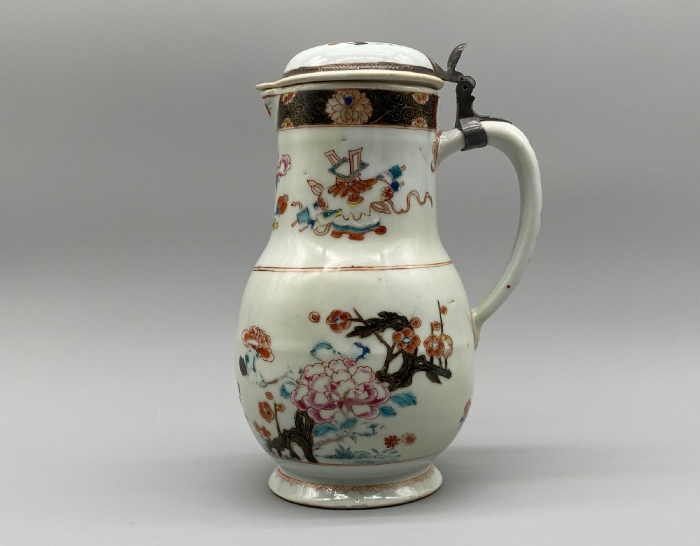 An 18th-century Chinese porcelain jug with hand-painted Famille Rose floral enamels, a metal-mounted lid, and a loop handle.