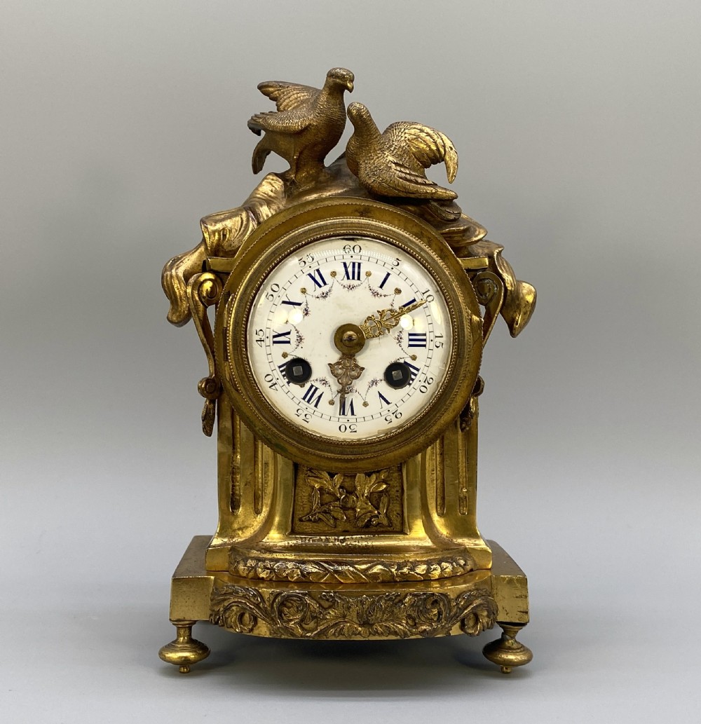 An antique French gilt bronze mantel clock surmounted by two sculpted doves, featuring an ornate pedestal base and a white enamel dial with Roman numerals. View II