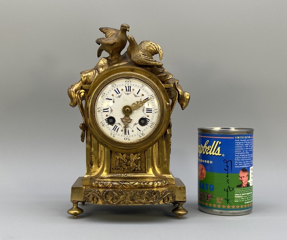 An antique French gilt bronze mantel clock surmounted by two sculpted doves, featuring an ornate pedestal base and a white enamel dial with Roman numerals. View III