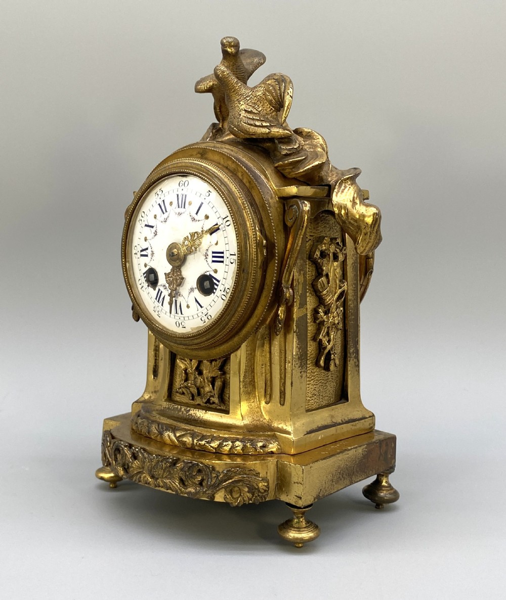 An antique French gilt bronze mantel clock surmounted by two sculpted doves, featuring an ornate pedestal base and a white enamel dial with Roman numerals. View V
