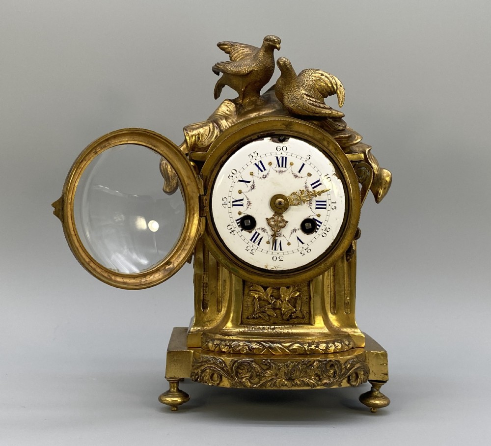 An antique French gilt bronze mantel clock surmounted by two sculpted doves, featuring an ornate pedestal base and a white enamel dial with Roman numerals. View IX