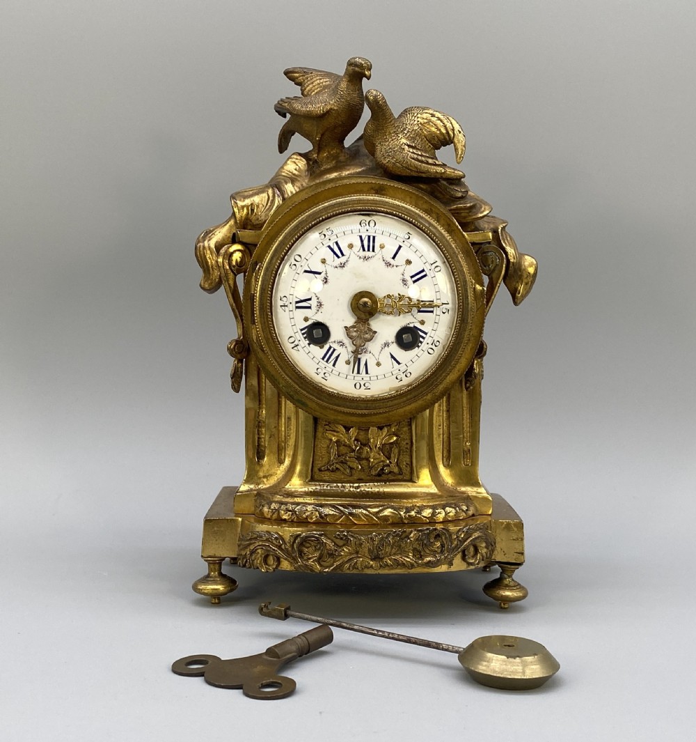 An antique French gilt bronze mantel clock surmounted by two sculpted doves, featuring an ornate pedestal base and a white enamel dial with Roman numerals. View X
