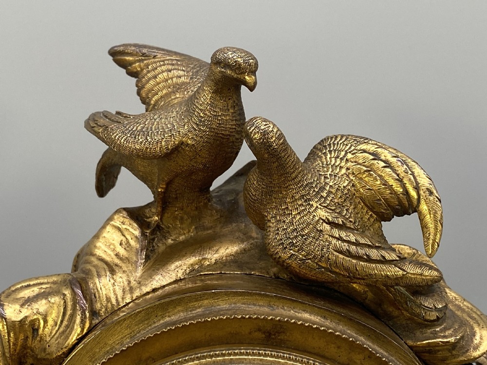 An antique French gilt bronze mantel clock surmounted by two sculpted doves, featuring an ornate pedestal base and a white enamel dial with Roman numerals. View XII