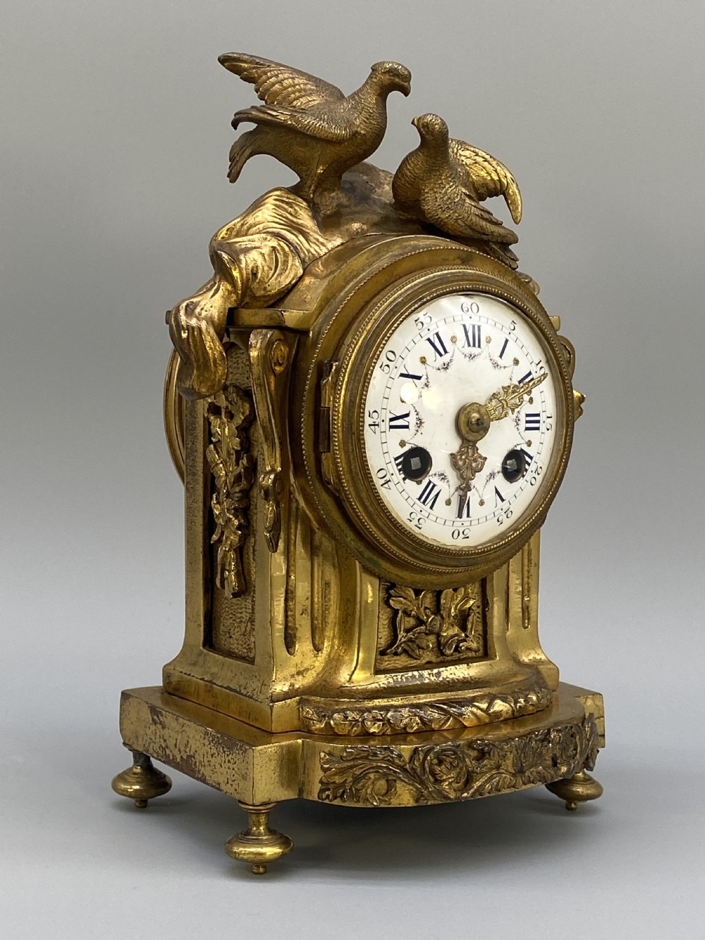 An antique French gilt bronze mantel clock surmounted by two sculpted doves, featuring an ornate pedestal base and a white enamel dial with Roman numerals. View XV