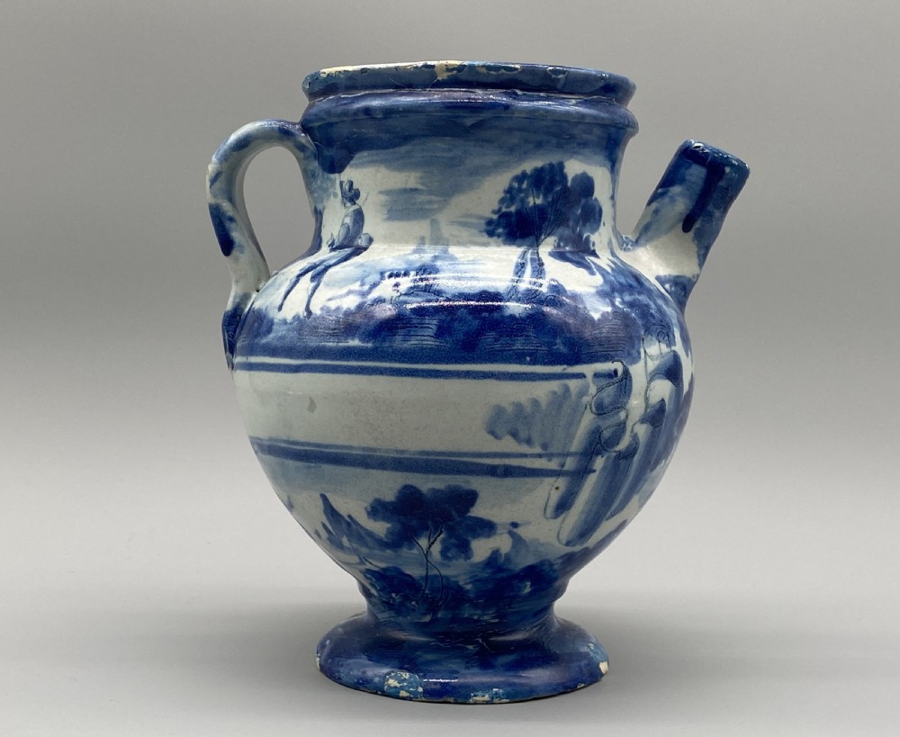 An 18th-century Italian Savona majolica wet drug jar featuring hand-painted cobalt blue landscapes, a single side handle, a raised spout, and a pedestal base.