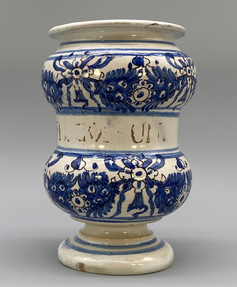 An 18th-century Italian majolica albarello pharmacy jar featuring a waisted silhouette with hand-painted blue floral patterns and central apothecary inscription.