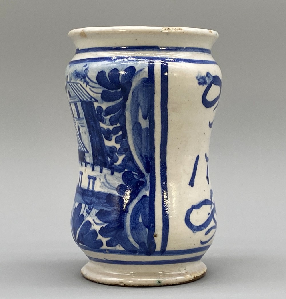 A cylindrical tin-glazed earthenware albarello jar with a waisted body featuring hand-painted blue scrollwork and the date 1709 on a cream-white background. View IV