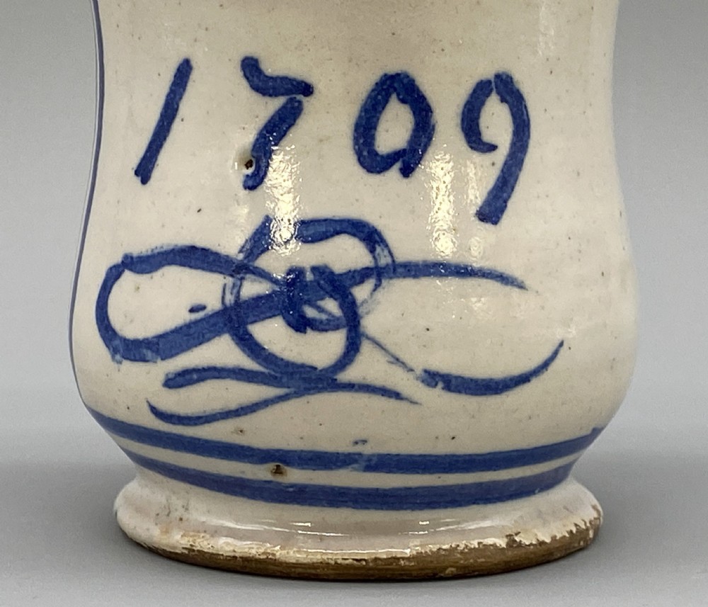 A cylindrical tin-glazed earthenware albarello jar with a waisted body featuring hand-painted blue scrollwork and the date 1709 on a cream-white background. View V