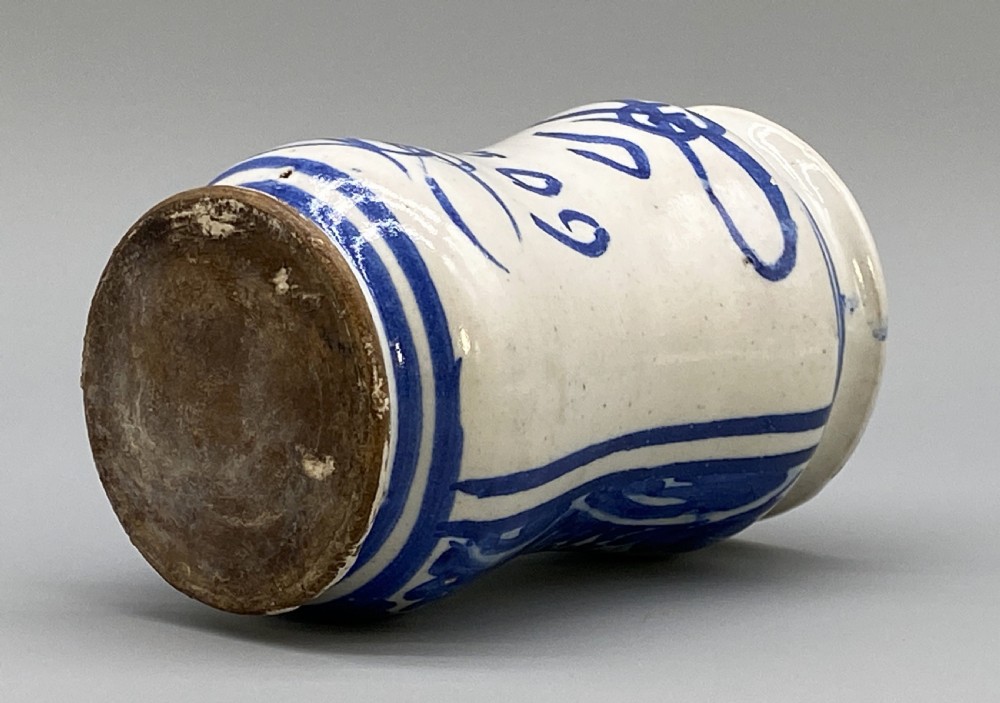 A cylindrical tin-glazed earthenware albarello jar with a waisted body featuring hand-painted blue scrollwork and the date 1709 on a cream-white background. View X