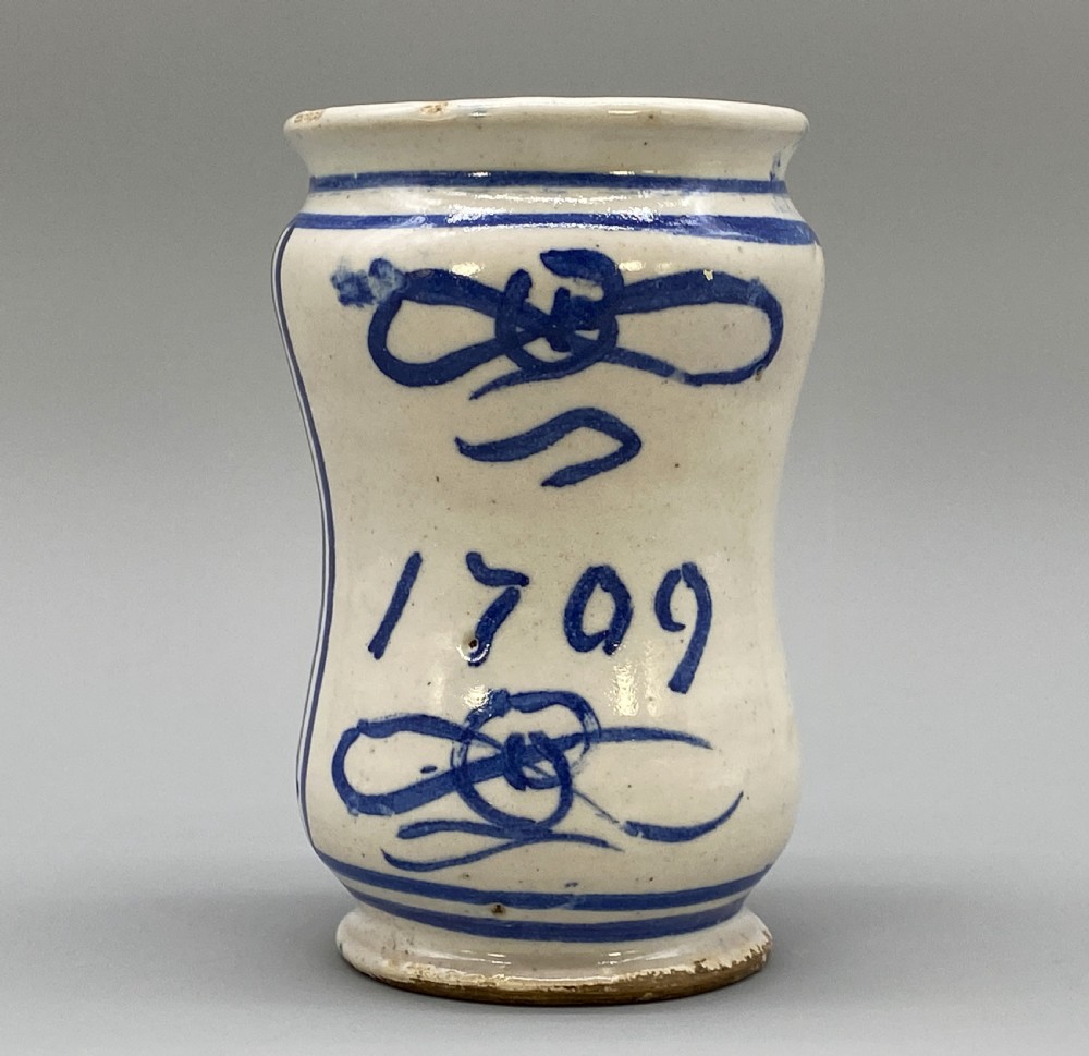 A cylindrical tin-glazed earthenware albarello jar with a waisted body featuring hand-painted blue scrollwork and the date 1709 on a cream-white background. View XIII