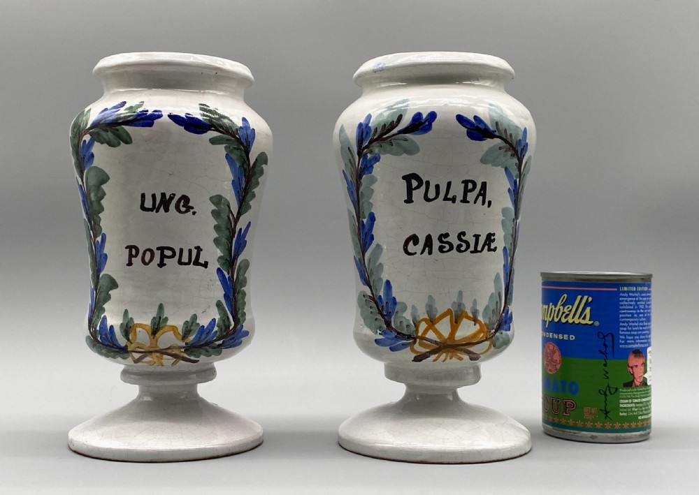 A pair of white tin-glazed ceramic albarello apothecary jars featuring hand-painted green foliage wreaths and Latin script on pedestal bases. View II