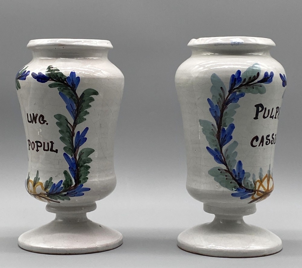 A pair of white tin-glazed ceramic albarello apothecary jars featuring hand-painted green foliage wreaths and Latin script on pedestal bases. View III