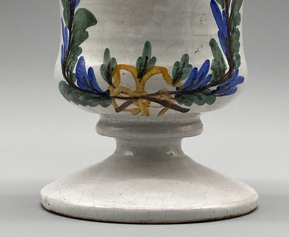 A pair of white tin-glazed ceramic albarello apothecary jars featuring hand-painted green foliage wreaths and Latin script on pedestal bases. View IX