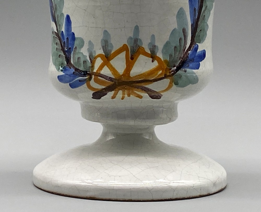 A pair of white tin-glazed ceramic albarello apothecary jars featuring hand-painted green foliage wreaths and Latin script on pedestal bases. View XIV