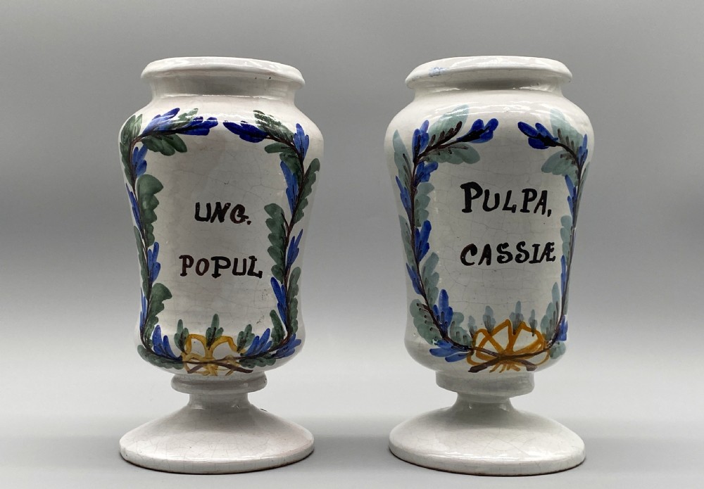 A pair of white tin-glazed ceramic albarello apothecary jars featuring hand-painted green foliage wreaths and Latin script on pedestal bases. View XVIII