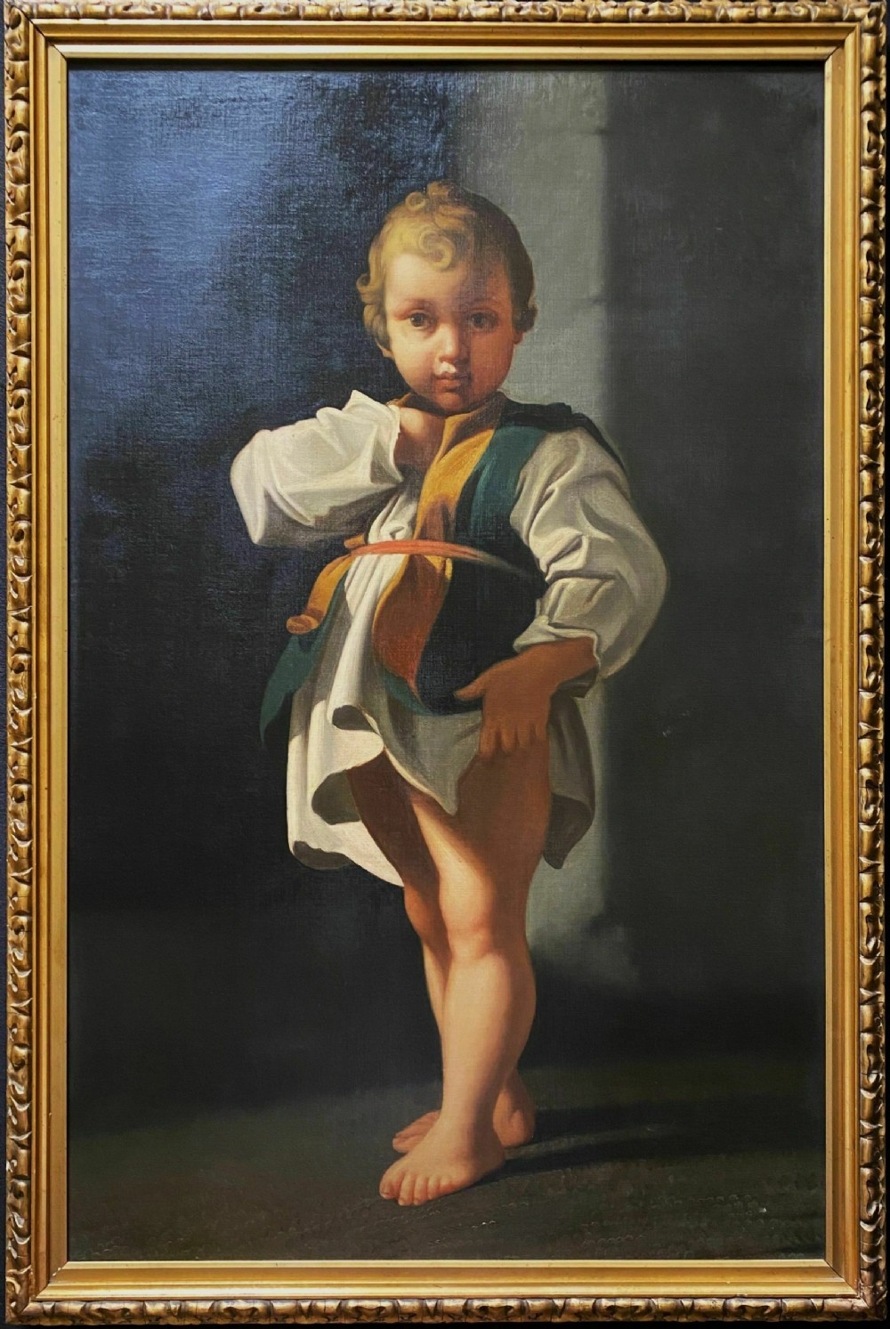 An eighteenth-century Baroque style oil painting on canvas depicting a standing young child dressed in a white tunic and green vest, set within a carved gilt frame.