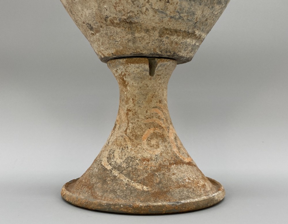 A painted terracotta vessel from the Tang Dynasty featuring a bulbous body, pointed finial lid, flared pedestal foot, and painted geometric scroll motifs in red and grey pigments. View X