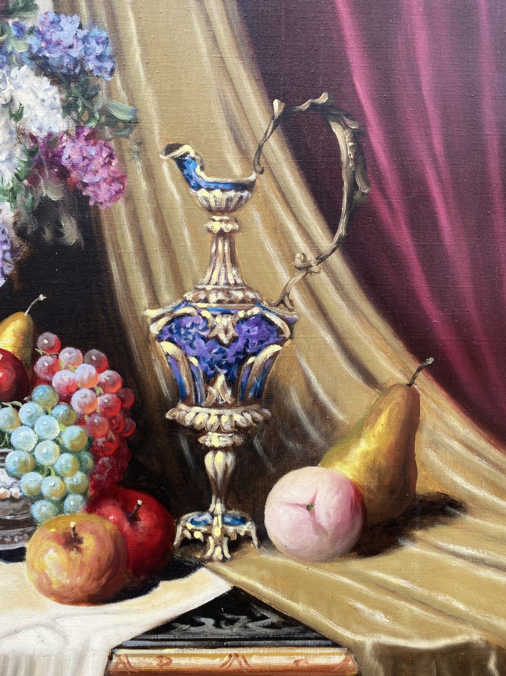 A framed oil painting on canvas depicting a table with fruit, lilacs in a central vase, silver ewers, and draped gold and burgundy fabrics. View IV