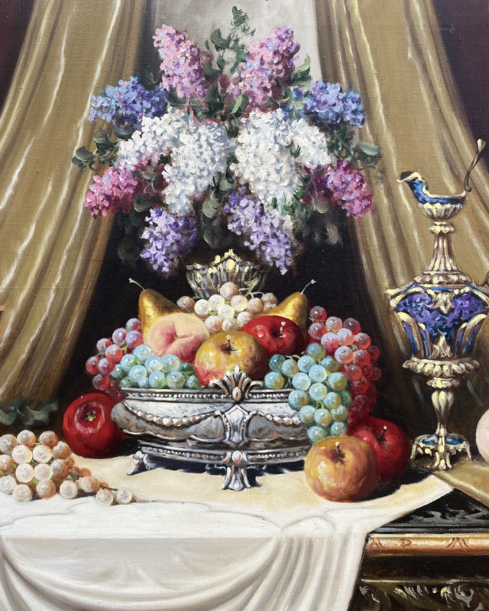 A framed oil painting on canvas depicting a table with fruit, lilacs in a central vase, silver ewers, and draped gold and burgundy fabrics. View V