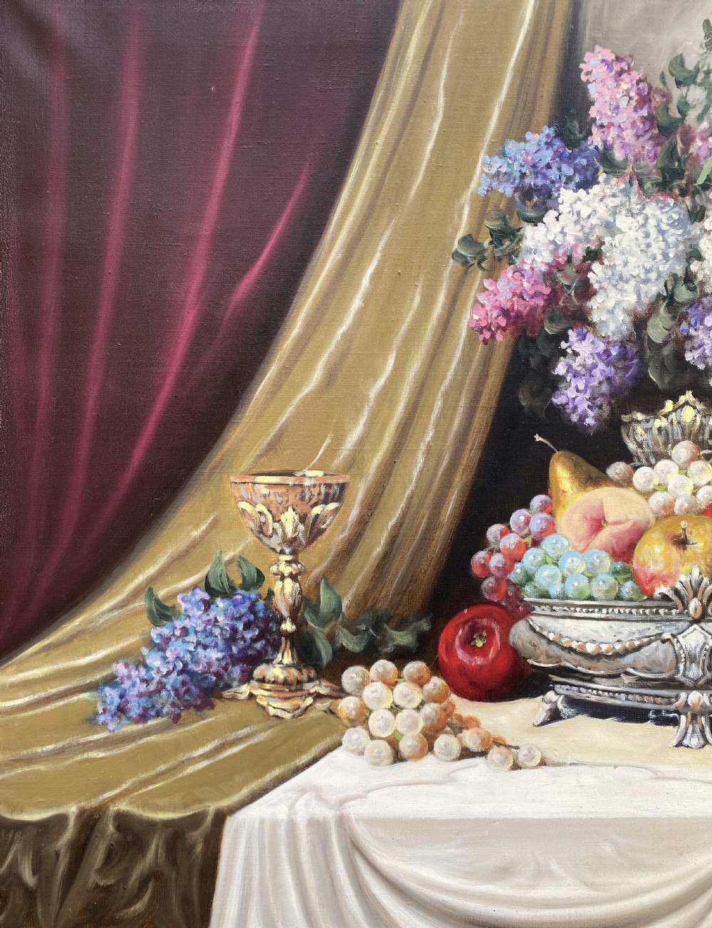 A framed oil painting on canvas depicting a table with fruit, lilacs in a central vase, silver ewers, and draped gold and burgundy fabrics. View VI