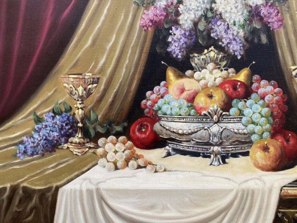 A framed oil painting on canvas depicting a table with fruit, lilacs in a central vase, silver ewers, and draped gold and burgundy fabrics. View VII