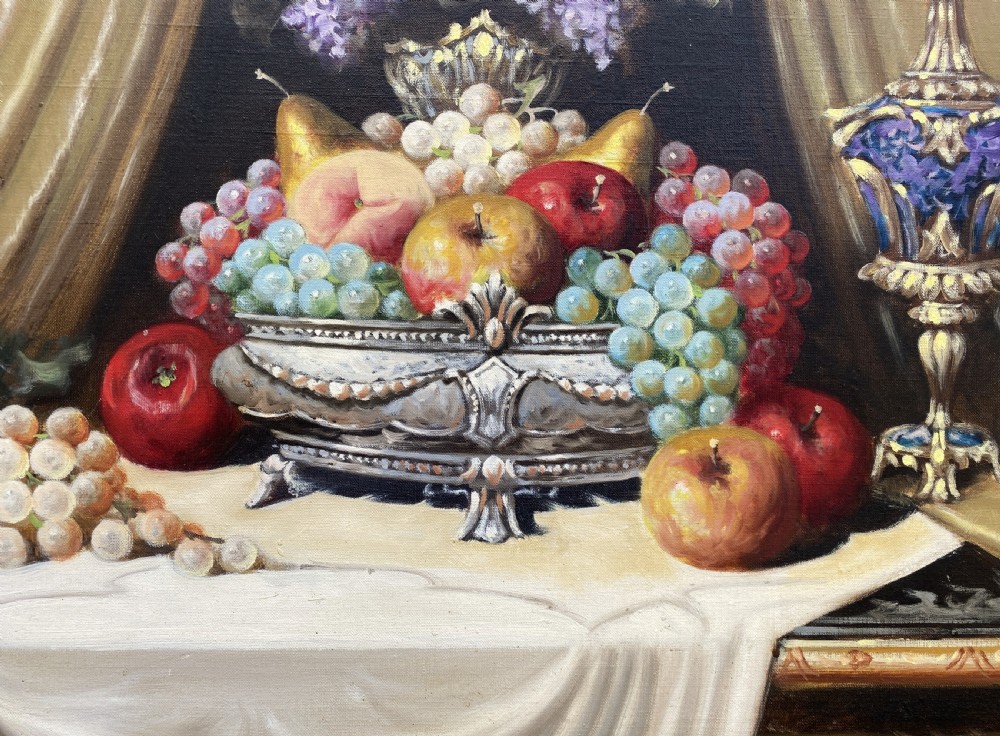 A framed oil painting on canvas depicting a table with fruit, lilacs in a central vase, silver ewers, and draped gold and burgundy fabrics. View VIII