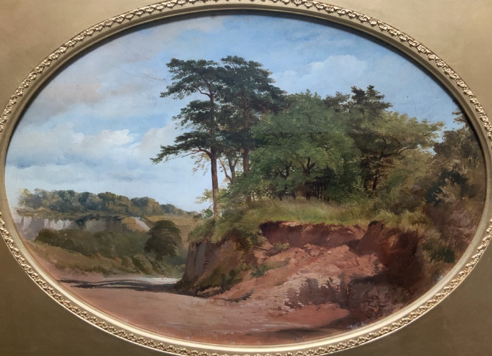 An oval 19th-century oil landscape painting depicting a wooded bank and open fields, set inside a rectangular gilded frame. View III