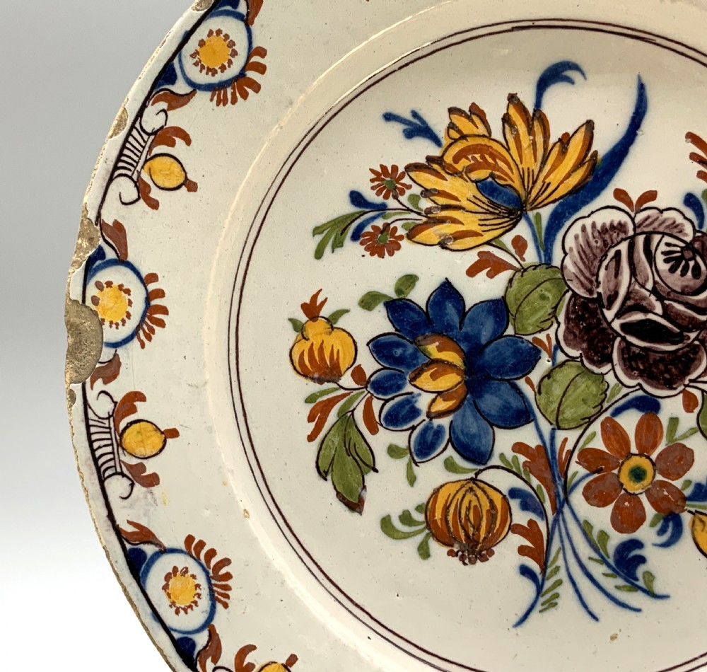 An 18th-century circular tin-glazed earthenware charger plate decorated with hand-painted floral motifs in manganese purple, cobalt blue, ochre yellow, and green. View VII