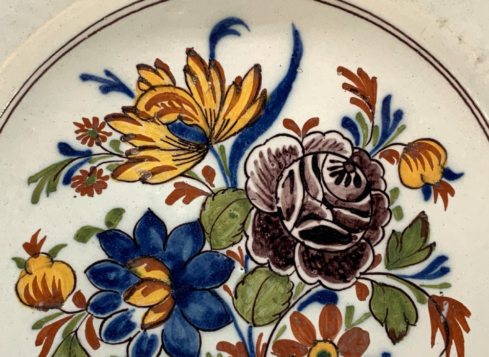 An 18th-century circular tin-glazed earthenware charger plate decorated with hand-painted floral motifs in manganese purple, cobalt blue, ochre yellow, and green. View IX