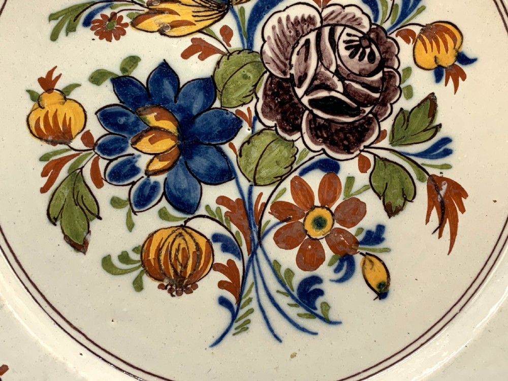 An 18th-century circular tin-glazed earthenware charger plate decorated with hand-painted floral motifs in manganese purple, cobalt blue, ochre yellow, and green. View X